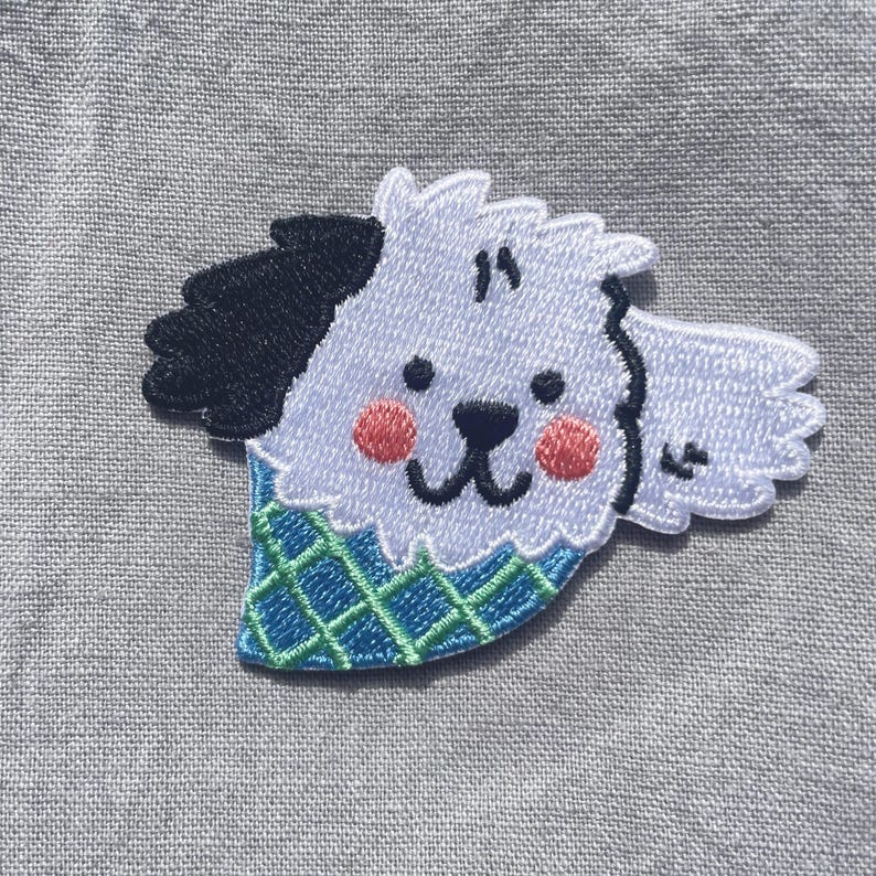 May include: An embroidered patch featuring a white dog with a black ear, pink cheeks, and a black nose and mouth. The dog is wearing a blue bandana with a green grid pattern. The embroidery is detailed with white, black, blue, green, and pink thread.