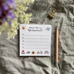 Op de afbeelding: Een wit notitieblok met de Duitse tekst "Meine kleinen Gl&uuml;cksmomente" en genummerde lijnen. Het notitieblok bevat kleine illustraties van een zon, vogel, bloem, hond, koffiekopje, fiets en regenboog. Een potlood en een gouden paperclip zijn ook aanwezig.