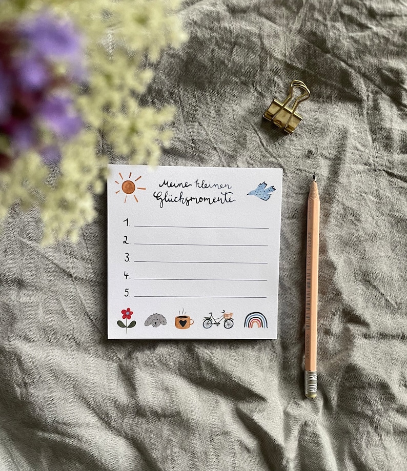 Op de afbeelding: Een wit notitieblok met de Duitse tekst "Meine kleinen Gl&uuml;cksmomente" en genummerde lijnen. Het notitieblok bevat kleine illustraties van een zon, vogel, bloem, hond, koffiekopje, fiets en regenboog. Een potlood en een gouden paperclip zijn ook aanwezig.