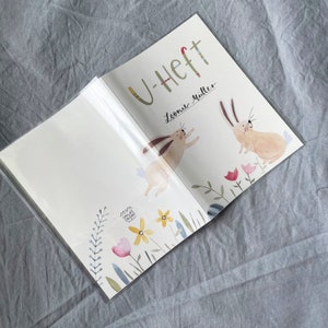 May include: A white book with a colorful cover featuring two bunnies and flowers. The book is titled "U-Heft" and has the name "Leonie M&uuml;ller" written on the cover.