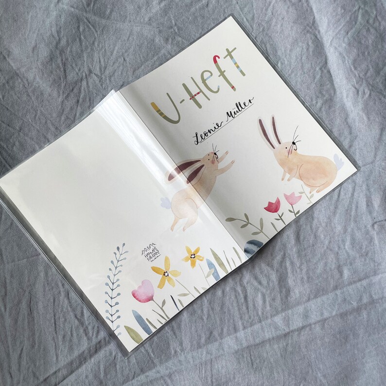 May include: A white book with a colorful cover featuring two bunnies and flowers. The book is titled "U-Heft" and has the name "Leonie M&uuml;ller" written on the cover.
