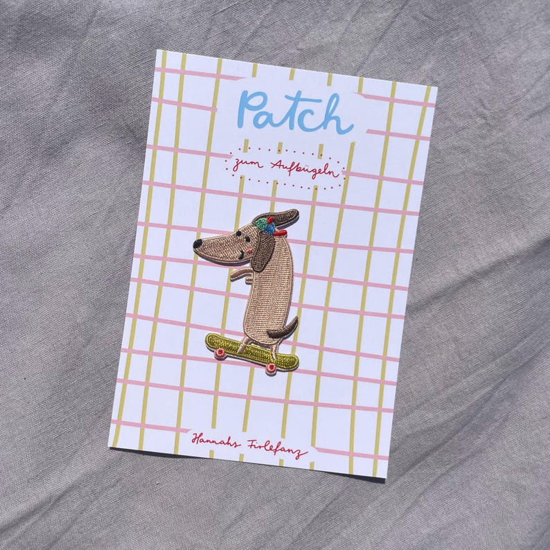 May include: Embroidered patch featuring a dachshund dog on a skateboard. The patch is on a card with a grid pattern and the word "Patch" in blue. The card also has the text "zum Aufb&uuml;geln" and "Hannahs Firlefanz".