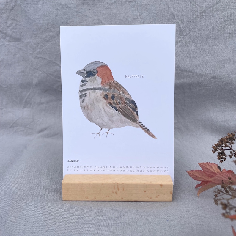 Puede incluir: Una p&aacute;gina de calendario vertical con una ilustraci&oacute;n en acuarela de un gorri&oacute;n com&uacute;n con plumaje marr&oacute;n, gris y negro. El texto "HAUSSPATZ" y "JANUAR" est&aacute; impreso en el papel blanco, sostenido en posici&oacute;n vertical por un bloque de madera.