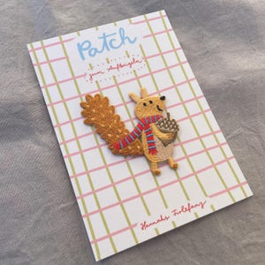 May include: Embroidered patch featuring a cartoon squirrel holding an acorn, wearing a red and blue striped scarf. The patch is on a card with a pink and green plaid pattern and the word "Patch" in blue.