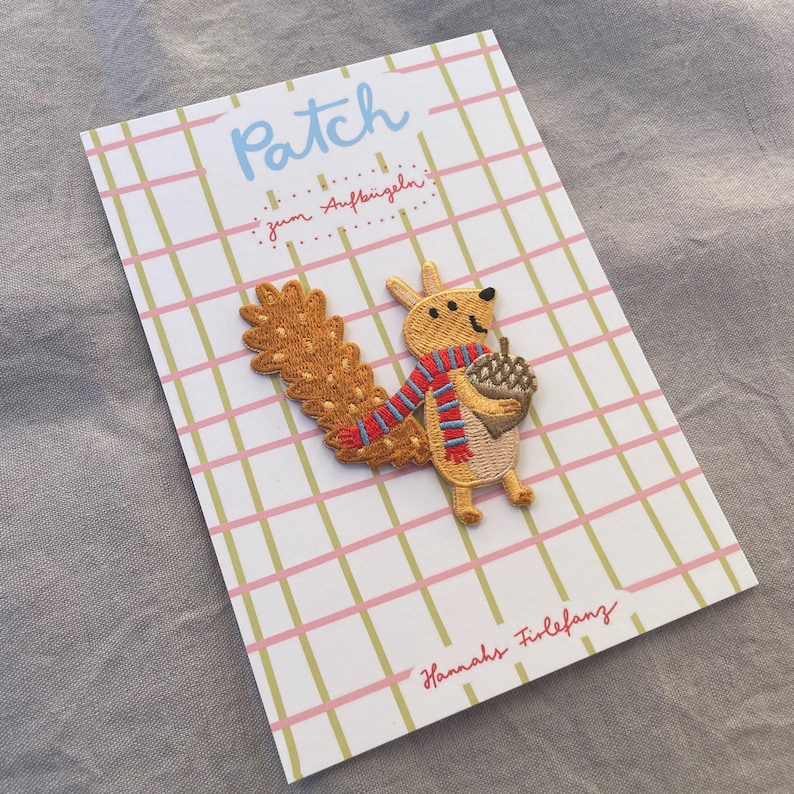 May include: Embroidered patch featuring a cartoon squirrel holding an acorn, wearing a red and blue striped scarf. The patch is on a card with a pink and green plaid pattern and the word "Patch" in blue.