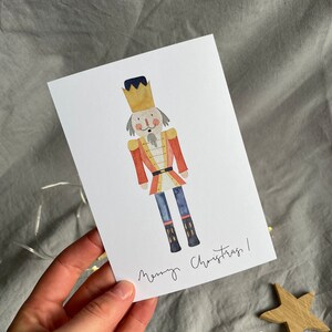 May include: A watercolor illustration of a nutcracker with a red and gold uniform, a blue hat, and a gold crown. The nutcracker is standing on a white background with the text "Merry Christmas!" written below.