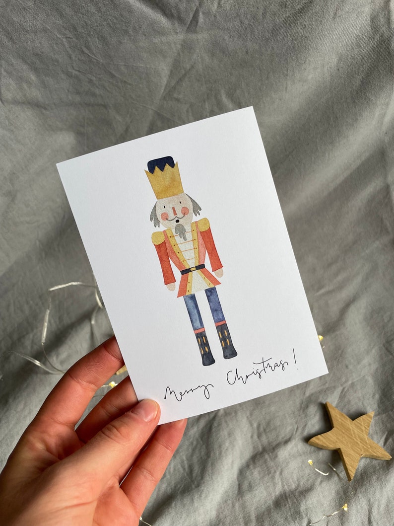 May include: A watercolor illustration of a nutcracker with a red and gold uniform, a blue hat, and a gold crown. The nutcracker is standing on a white background with the text "Merry Christmas!" written below.