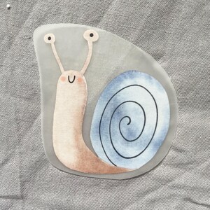 Pu&ograve; includere: Un adesivo stravagante con una lumaca dei cartoni animati con un corpo rosa chiaro e un guscio a spirale blu e bianco. La lumaca ha lunghe antenne con occhi neri e una faccia sorridente. L'adesivo &egrave; su sfondo grigio.