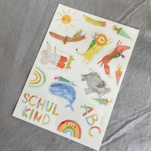 Pu&ograve; includere: Un foglio di adesivi colorati con illustrazioni di animali stravaganti, tra cui un sole, un bassotto, un leone e una volpe. Gli adesivi includono anche un arcobaleno, una balena e le parole "SCHUL KIND" e "ABC".