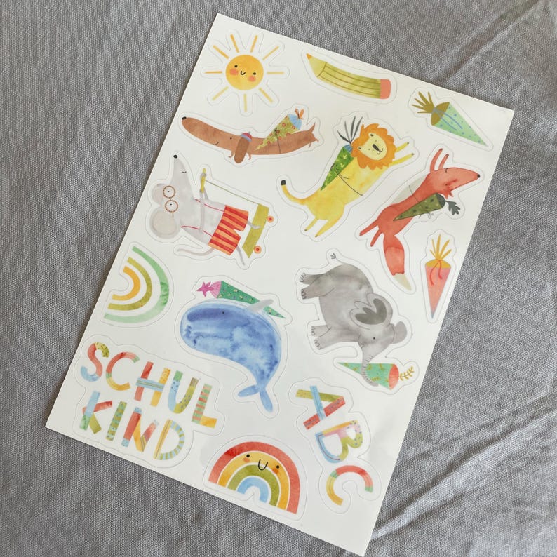 Pu&ograve; includere: Un foglio di adesivi colorati con illustrazioni di animali stravaganti, tra cui un sole, un bassotto, un leone e una volpe. Gli adesivi includono anche un arcobaleno, una balena e le parole "SCHUL KIND" e "ABC".