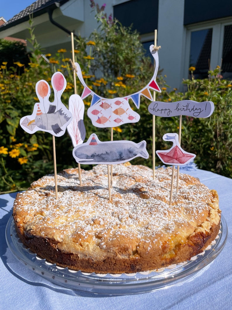 May include: A round cake with a light brown crust and a white powdered sugar topping. The cake is decorated with colorful paper cutouts of fish, a whale, a bird, and a banner that says "happy birthday!"