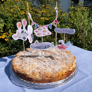 May include: A round cake with a white crumb topping and a variety of colorful decorations, including a whale, fish, and a banner that says "Happy Birthday!" The cake is on a glass plate and a blue tablecloth.