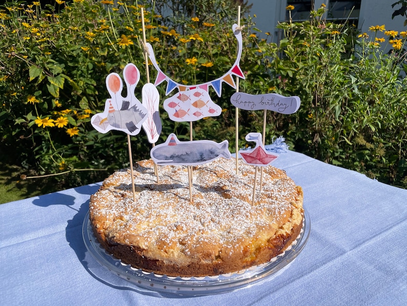 May include: A round cake with a white crumb topping and a variety of colorful decorations, including a whale, fish, and a banner that says "Happy Birthday!" The cake is on a glass plate and a blue tablecloth.