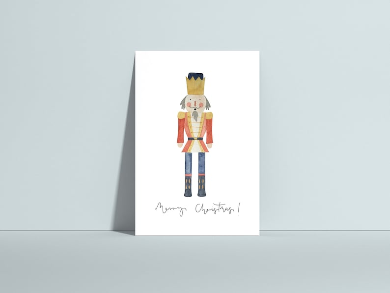 May include: A watercolor illustration of a nutcracker soldier wearing a red and blue uniform with a gold crown. The text "Merry Christmas!" is written below the nutcracker.