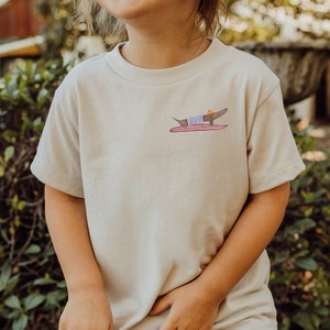 May include: A light beige t-shirt featuring a graphic of a dachshund wearing a striped shirt and a hat, riding a surfboard.