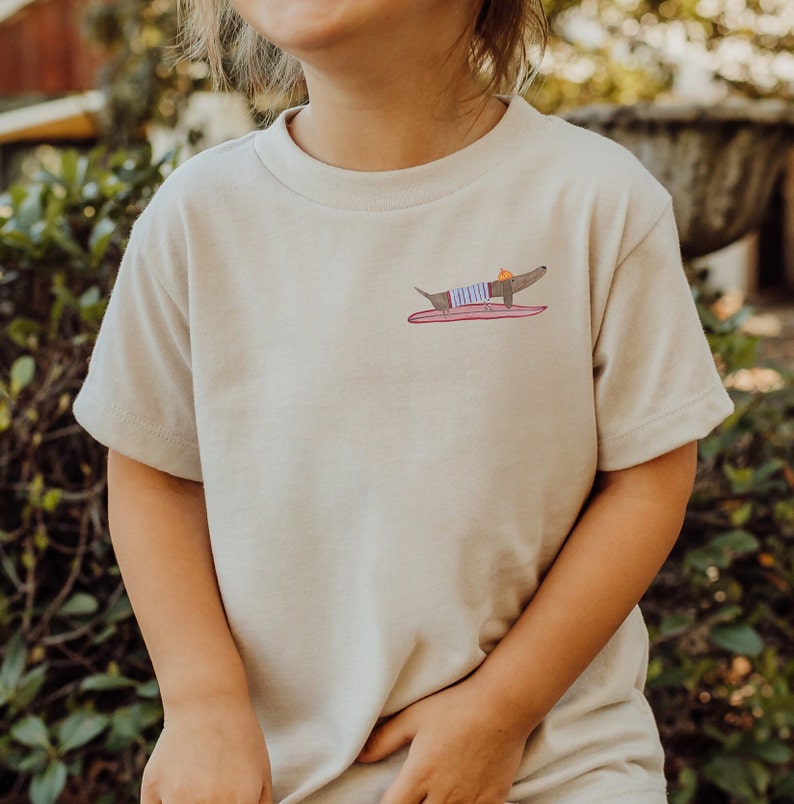 May include: A light beige t-shirt featuring a graphic of a dachshund wearing a striped shirt and a hat, riding a surfboard.