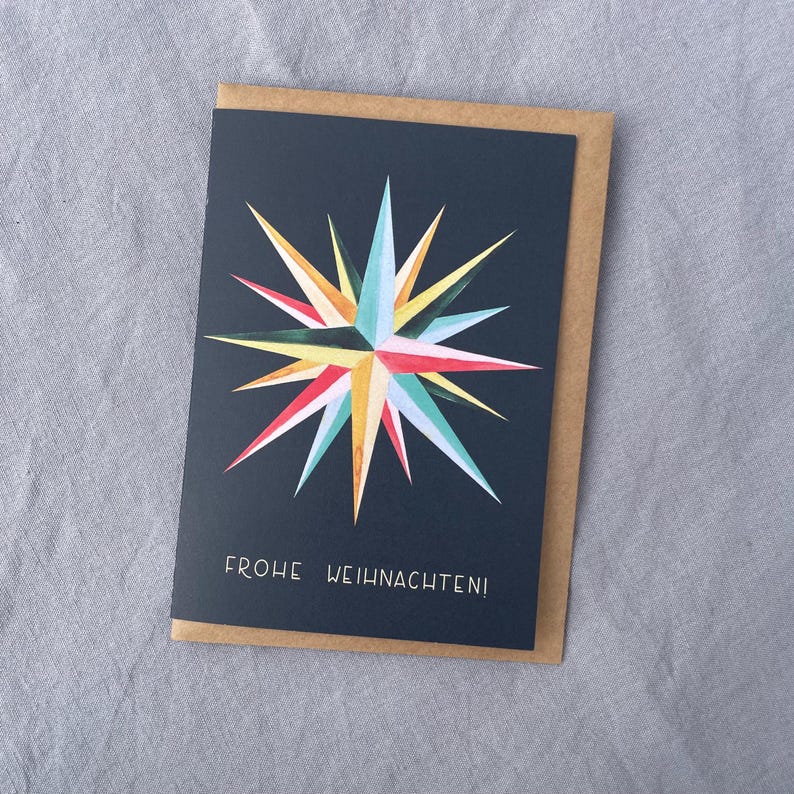 May include: A greeting card with a navy blue background and a colorful starburst design in red, yellow, turquoise, and white. The card reads "FROHE WEIHNACHTEN!" in white lettering and is inside a brown envelope.
