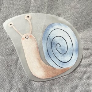 Pu&ograve; includere: Un adesivo stravagante con una lumaca sorridente con un corpo marrone chiaro e un guscio blu e grigio. La lumaca ha lunghe antenne con punte nere. L'adesivo &egrave; su uno sfondo trasparente.