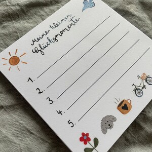 Op de afbeelding: Een witte notitieblok met de Duitse tekst "Meine kleinen Gl&uuml;cksmomente" en vijf genummerde lijnen. Het notitieblok bevat illustraties van een zon, vogel, fiets, koffiekopje, hond en bloem. Het notitieblok staat schuin op een grijze ondergrond.