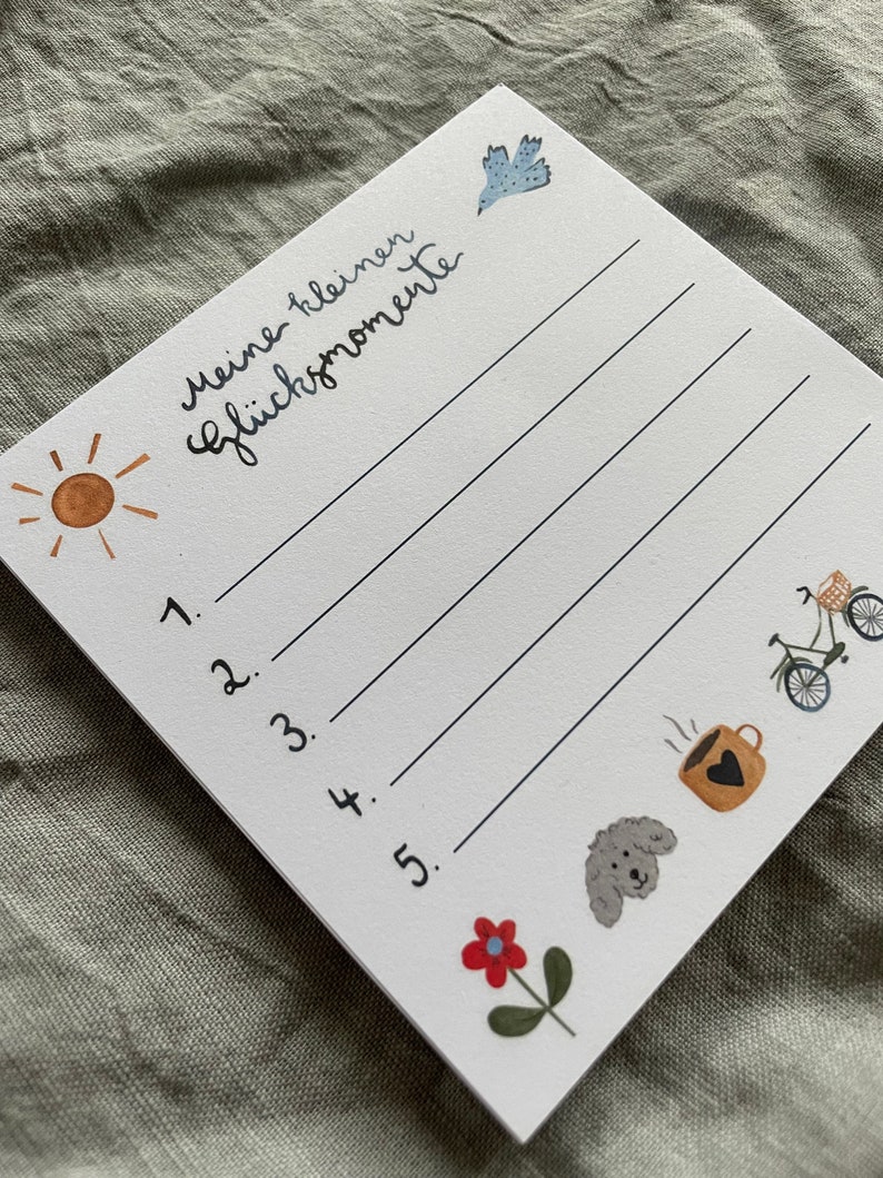 Op de afbeelding: Een witte notitieblok met de Duitse tekst "Meine kleinen Gl&uuml;cksmomente" en vijf genummerde lijnen. Het notitieblok bevat illustraties van een zon, vogel, fiets, koffiekopje, hond en bloem. Het notitieblok staat schuin op een grijze ondergrond.