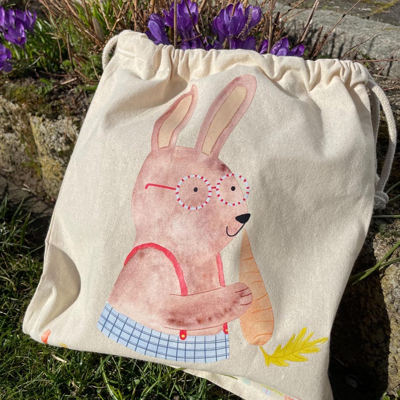 May include: A beige canvas drawstring bag with a cartoon illustration of a brown bunny wearing red and white glasses and holding a carrot. The bunny is wearing a blue and white plaid shirt and red suspenders.