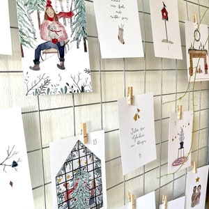 Puede incluir: Una colecci&oacute;n de tarjetas navide&ntilde;as ilustradas, sujetas a una rejilla met&aacute;lica con pinzas de madera. Las tarjetas presentan escenas invernales, decoraciones festivas y texto navide&ntilde;o. Las tarjetas est&aacute;n numeradas e incluyen im&aacute;genes de una escena de trineo, una casita para p&aacute;jaros y un &aacute;rbol de Navidad.
