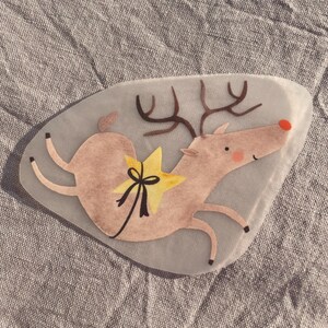 May include: A whimsical illustration of a reindeer with a yellow star and black bow. The reindeer is light brown with a red nose and brown antlers. The image has a watercolor style and is set against a gray background.
