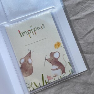 May include: A rectangular vaccination record booklet cover featuring a cream-colored background with two brown watercolor mice, green grass, a pink flower, and a yellow dandelion. The text Impfpass is printed in multicolored letters at the top.