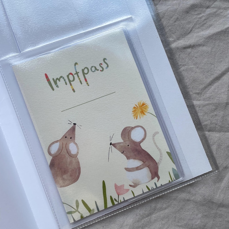 May include: A rectangular vaccination record booklet cover featuring a cream-colored background with two brown watercolor mice, green grass, a pink flower, and a yellow dandelion. The text Impfpass is printed in multicolored letters at the top.