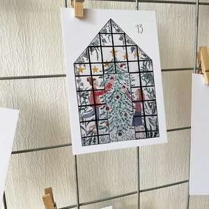 Puede incluir: Tarjeta navide&ntilde;a con un dise&ntilde;o en forma de casa, que presenta un &aacute;rbol de Navidad y dos figuras decor&aacute;ndolo. La tarjeta est&aacute; sujeta por una pinza de madera en una rejilla met&aacute;lica. El n&uacute;mero "13" es visible.