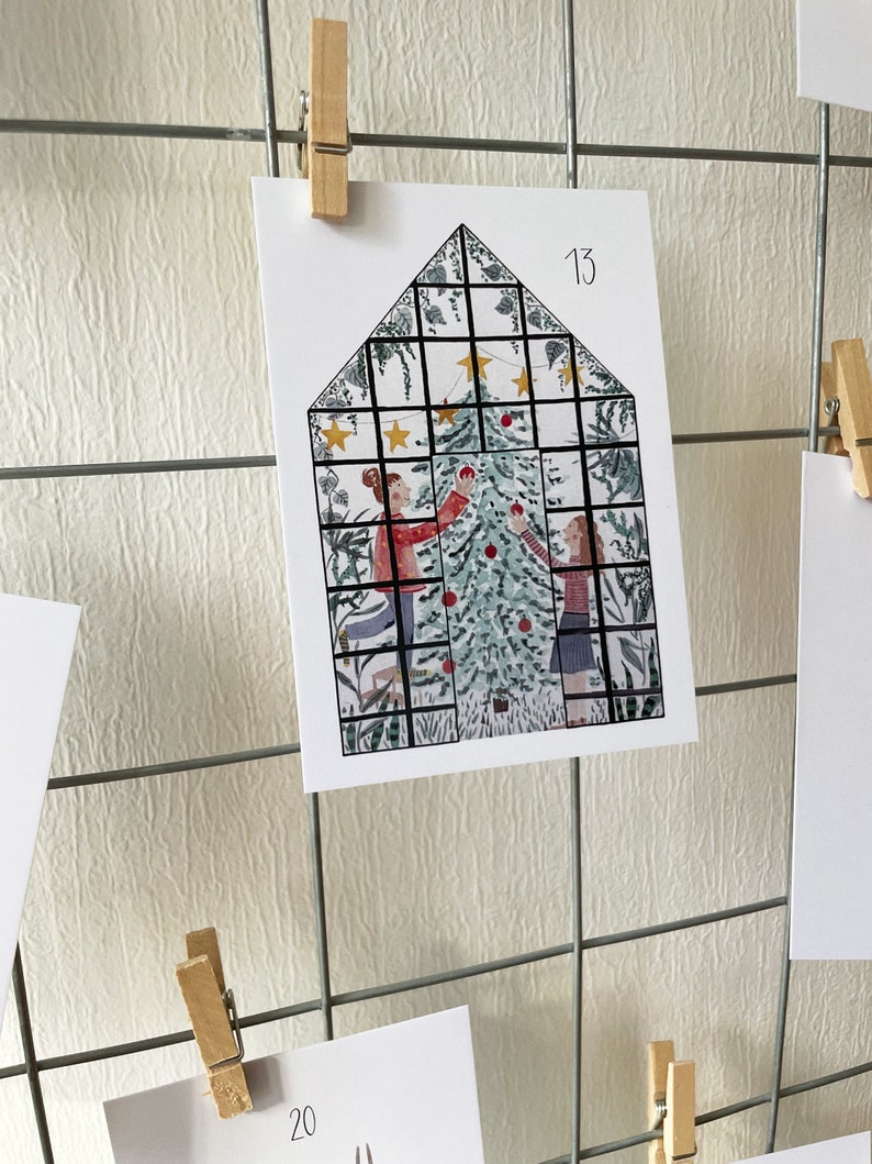 Puede incluir: Tarjeta navide&ntilde;a con un dise&ntilde;o en forma de casa, que presenta un &aacute;rbol de Navidad y dos figuras decor&aacute;ndolo. La tarjeta est&aacute; sujeta por una pinza de madera en una rejilla met&aacute;lica. El n&uacute;mero "13" es visible.