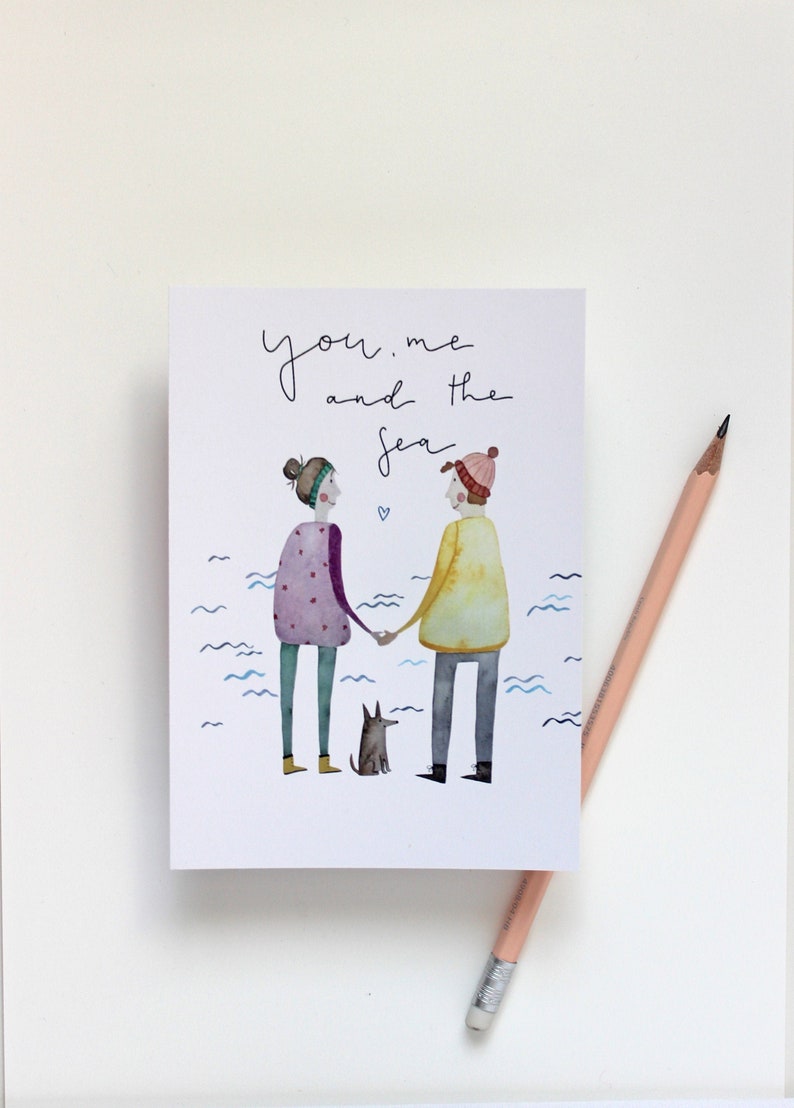 Puede incluir: Una tarjeta de felicitaci&oacute;n con el texto "you, me and the sea" sobre una ilustraci&oacute;n de dos figuras tomadas de la mano, un perro peque&ntilde;o y l&iacute;neas onduladas azules. Un l&aacute;piz descansa sobre la superficie blanca.
