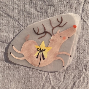 May include: A whimsical, light brown reindeer illustration with a yellow star and black bow. The reindeer has brown antlers, a red nose, and is set against a light gray background. The design is likely for a holiday-themed craft or decoration.
