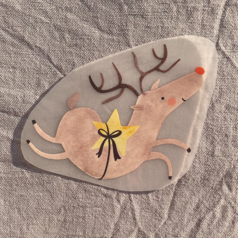 May include: A whimsical, light brown reindeer illustration with a yellow star and black bow. The reindeer has brown antlers, a red nose, and is set against a light gray background. The design is likely for a holiday-themed craft or decoration.