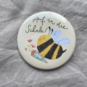 May include: A round button featuring a cartoon bee with yellow and black stripes, light blue wings, and a small floral cone. The text "Auf in die Schule!" is written in black script above the bee on a cream-colored background.