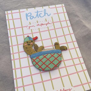 May include: Embroidered patch of a cartoon pickle in a baseball cap, relaxing in a blue and green basket with a red diamond pattern. The patch is on a card with the word "Patch" and text in German.