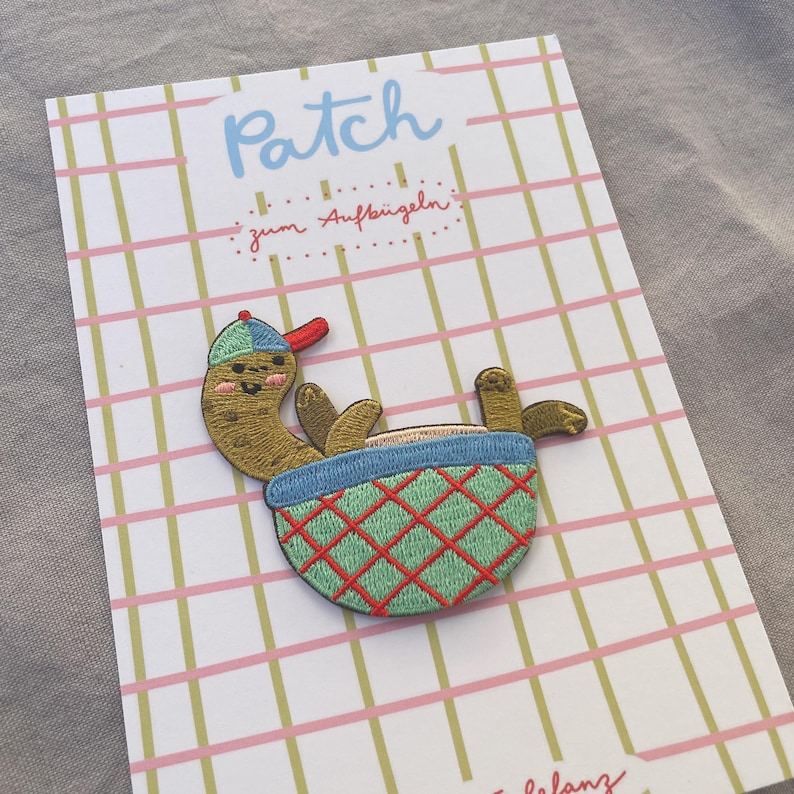 May include: Embroidered patch of a cartoon pickle in a baseball cap, relaxing in a blue and green basket with a red diamond pattern. The patch is on a card with the word "Patch" and text in German.