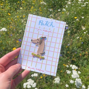 May include: A patch featuring a dachshund dog riding a skateboard. The dog is brown with a colorful headband. The patch is on a card with the word "Patch" and the text "zum Aufb&uuml;geln". The card has a pink and yellow grid pattern.