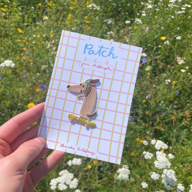 May include: A patch featuring a dachshund dog riding a skateboard. The dog is brown with a colorful headband. The patch is on a card with the word "Patch" and the text "zum Aufb&uuml;geln". The card has a pink and yellow grid pattern.