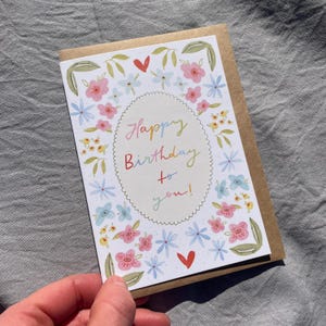 Może przedstawiać: Kwiatowa kartka urodzinowa z napisem "Happy Birthday to you!" w kolorowym skrypcie. Karta zawiera r&oacute;żowe, niebieskie i ż&oacute;łte kwiaty oraz zielone liście. Karta jest trzymana na szarym tle.