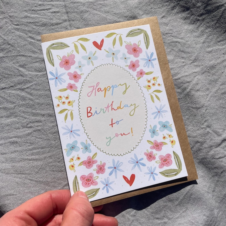 Może przedstawiać: Kwiatowa kartka urodzinowa z napisem "Happy Birthday to you!" w kolorowym skrypcie. Karta zawiera r&oacute;żowe, niebieskie i ż&oacute;łte kwiaty oraz zielone liście. Karta jest trzymana na szarym tle.