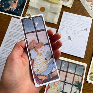 Op de afbeelding: Waterverf bladwijzer met een persoon die leest bij een raam met een hond. De bladwijzer heeft een touwtje om op te hangen. Andere items zijn vogelstickers, een met sterren gevulde illustratie en een vogelgids.