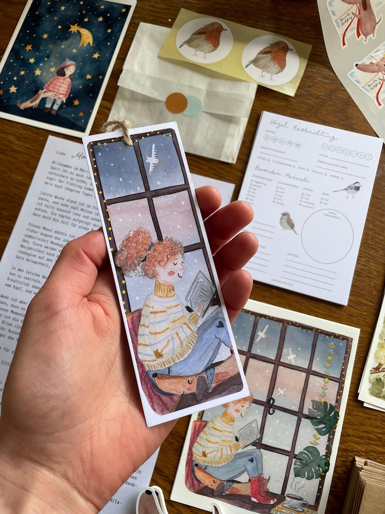Op de afbeelding: Waterverf bladwijzer met een persoon die leest bij een raam met een hond. De bladwijzer heeft een touwtje om op te hangen. Andere items zijn vogelstickers, een met sterren gevulde illustratie en een vogelgids.