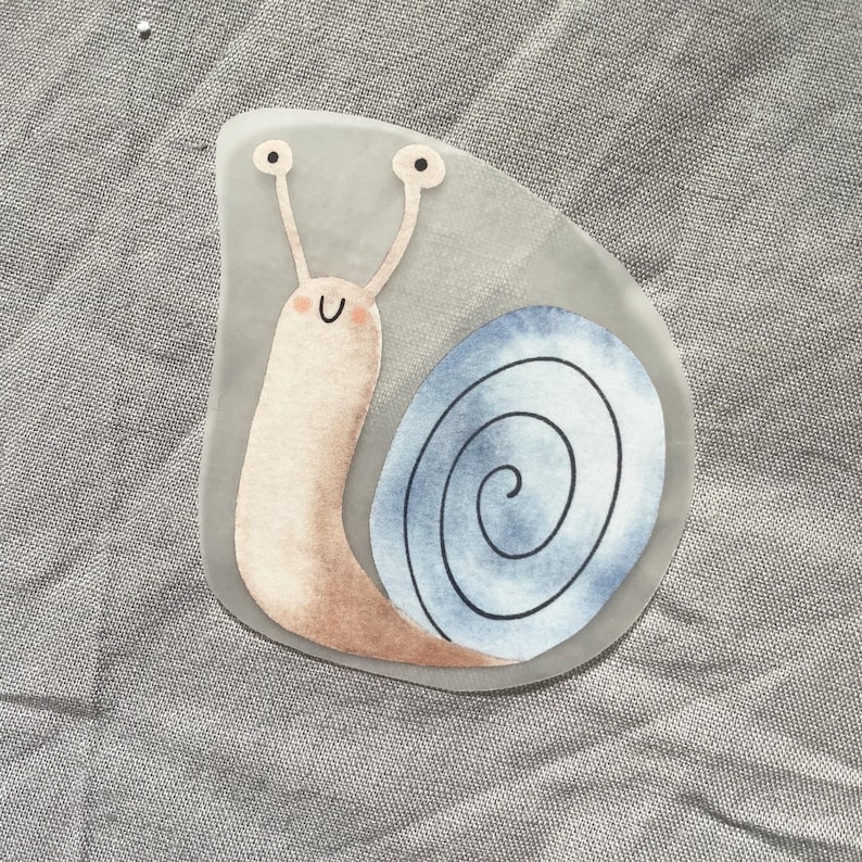 Pu&ograve; includere: Adesivo a forma di lumaca con corpo rosa chiaro e conchiglia a spirale blu e bianca. La lumaca ha due antenne con puntini neri e un viso sorridente. Sfondo grigio.