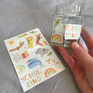 Pu&ograve; includere: Un barattolo di vetro trasparente con adesivi colorati, tra cui un arcobaleno, una carota e le parole "SCHUL KIND". Un foglio di adesivi con illustrazioni di animali e le lettere "ABC" &egrave; accanto al barattolo. Gli adesivi sono in stile acquerello.