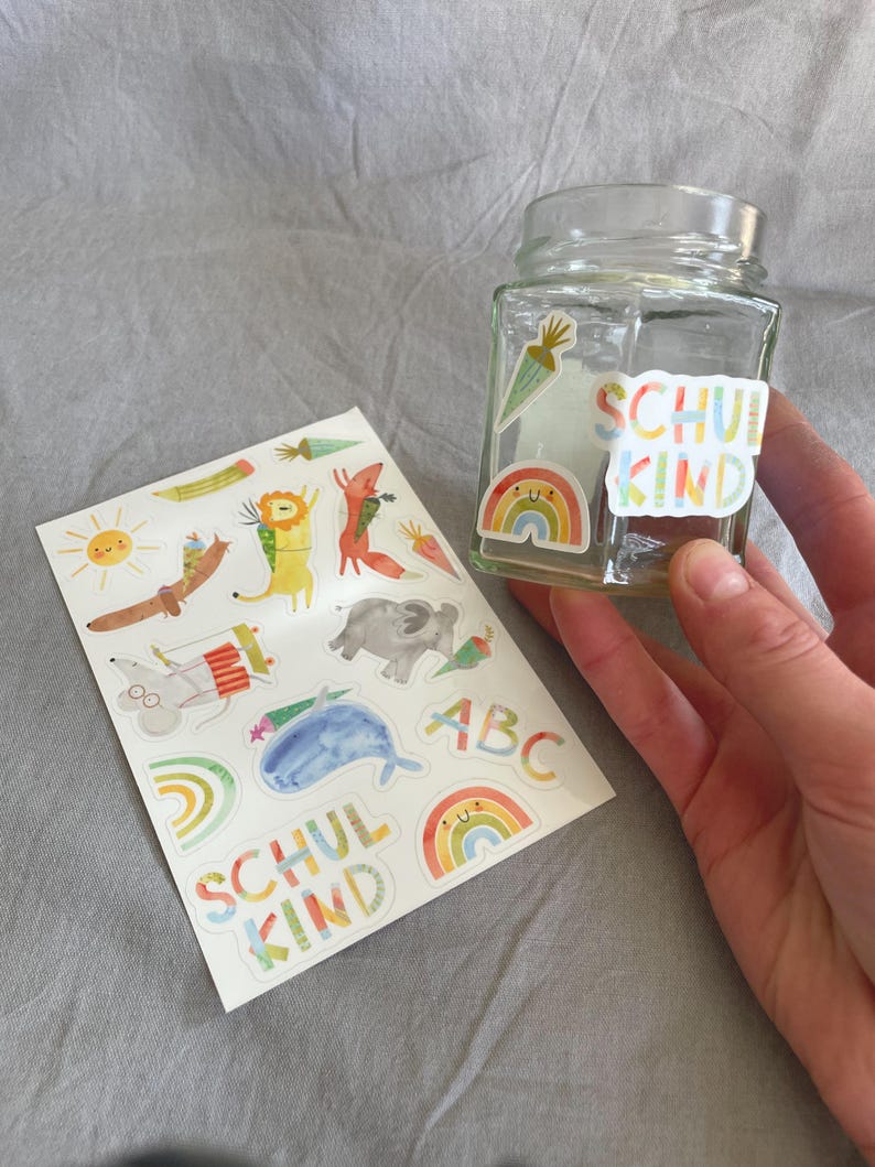 Pu&ograve; includere: Un barattolo di vetro trasparente con adesivi colorati, tra cui un arcobaleno, una carota e le parole "SCHUL KIND". Un foglio di adesivi con illustrazioni di animali e le lettere "ABC" &egrave; accanto al barattolo. Gli adesivi sono in stile acquerello.