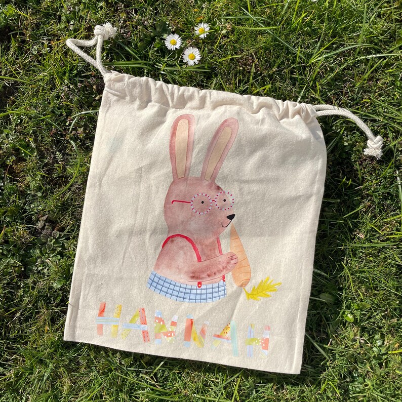May include: A beige drawstring bag with a bunny rabbit illustration. The bunny is wearing glasses and holding a carrot. The bag is personalized with the name "Hannah".