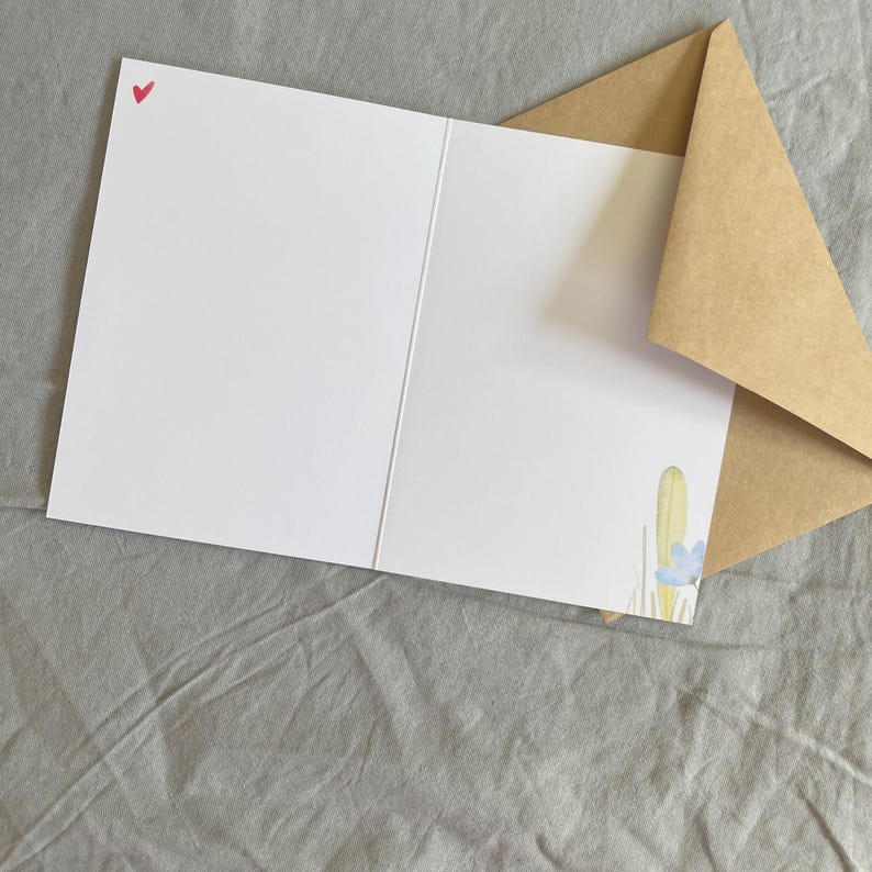 May include: A white greeting card with a small red heart in the top left corner. The card is open, showing a watercolour illustration of a yellow cactus and blue flower. A brown envelope is partially visible.