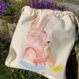 May include: A white canvas drawstring bag with a cartoon bunny wearing red glasses and holding a carrot. The bunny is wearing a blue and white checkered shirt.