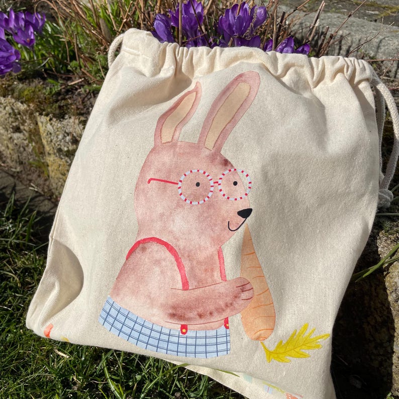 May include: A white canvas drawstring bag with a cartoon bunny wearing red glasses and holding a carrot. The bunny is wearing a blue and white checkered shirt.
